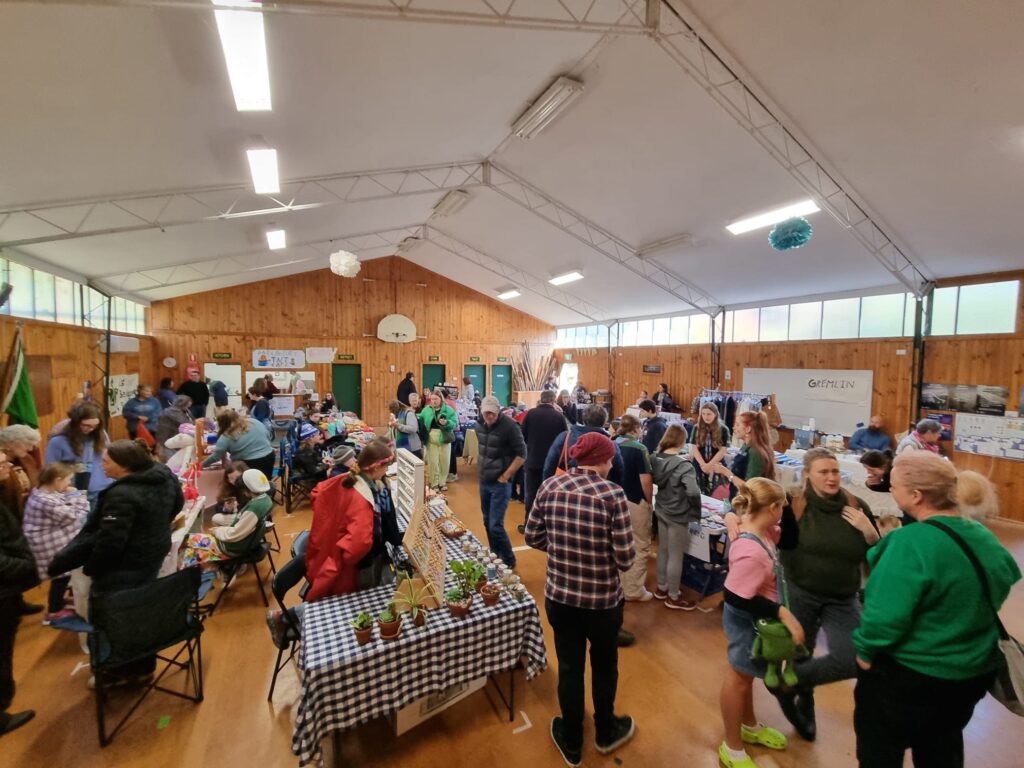 Photograph of the inside of the 1st Belgrave South Scout Hall during the Dandenong Ranges Youth Market. A long room with a white ceiling, old style metal internal roof frames, wooden walls and floor. A busy market with stalls and people chatting and browsing.