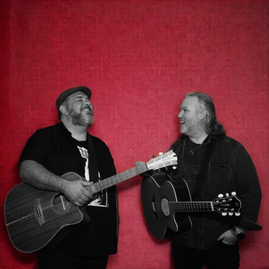 Black and white photo of Lloyd Spiegel and Charlie A’Court on top of red background. Holding guitars with happy expressions.