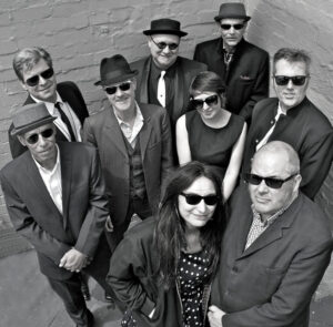 Black and white photo of the band Ska Vendors. Taken from above, looking down at the band. All members are smartly dressed in suits or dresses. All wearing sunglasses. Three members wearing hats.