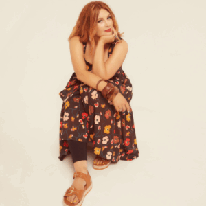 Studio colour portrait of Clare. Cream background. Sitting on a concealed stool. Wearing sandals and long dress with a flower design. Hand in chin with elbow and other arm resting on top of legs. Relaxed look on her face with a slight smile. Red lipstick.