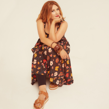 Studio colour portrait of Clare. Cream background. Sitting on a concealed stool. Wearing sandals and long dress with a flower design. Hand in chin with elbow and other arm resting on top of legs. Relaxed look on her face with a slight smile. Red lipstick.
