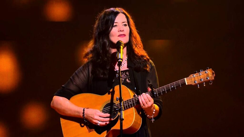 Colour photograph of Marcia Howard playing a live gig. Strumming an acoustic guitar in front of a microphone stand while looking out into the crowd. Dressed in black.