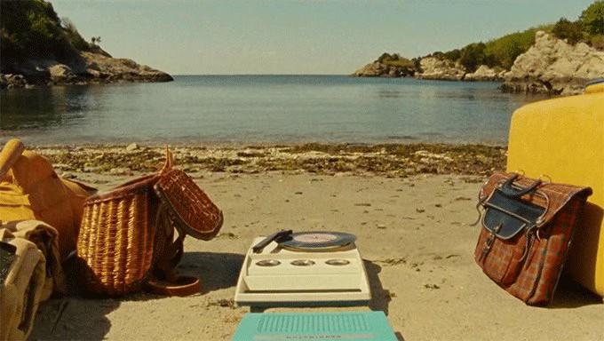 Film still from the movie Moonrise Kingdom. Portable record player, bags, basket and portable record player on a beach with the ocean in the background.