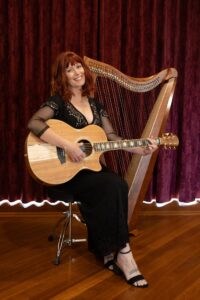 Colour photo of Beck Sian sitting on a stool holding a guitar with a harp behind her.